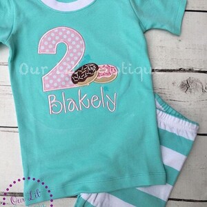 May include: A turquoise short-sleeved shirt with a pink and white polka dot number two and two donuts. The shirt says "Blakely" underneath the number. The shirt is paired with white and turquoise striped pants.