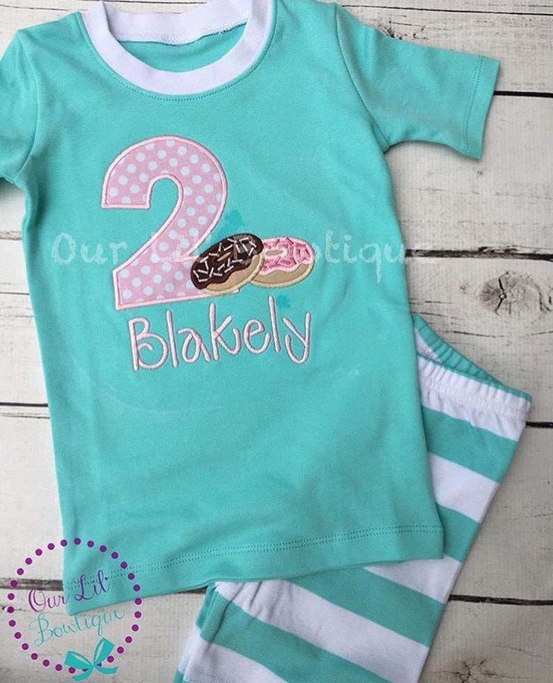 May include: A turquoise short-sleeved shirt with a pink and white polka dot number two and two donuts. The shirt says "Blakely" underneath the number. The shirt is paired with white and turquoise striped pants.