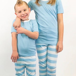 K&ouml;nnte beinhalten: Zwei Kinder in passenden hellblauen Pyjamas. Die Oberteile haben einen wei&szlig;en Besatz. Die Schlafanzughosen sind hellblau und wei&szlig; gestreift. Ein Kind hat eine hellblaue Schleife im Haar.