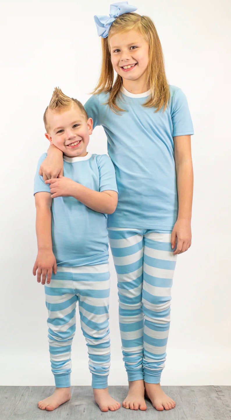 K&ouml;nnte beinhalten: Zwei Kinder in passenden hellblauen Pyjamas. Die Oberteile haben einen wei&szlig;en Besatz. Die Schlafanzughosen sind hellblau und wei&szlig; gestreift. Ein Kind hat eine hellblaue Schleife im Haar.