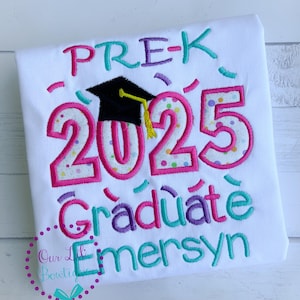 May include: White t-shirt with pink, blue, and purple embroidery that says "Pre-K 2025 Graduate" with a black graduation cap and the name "Emersyn" underneath.
