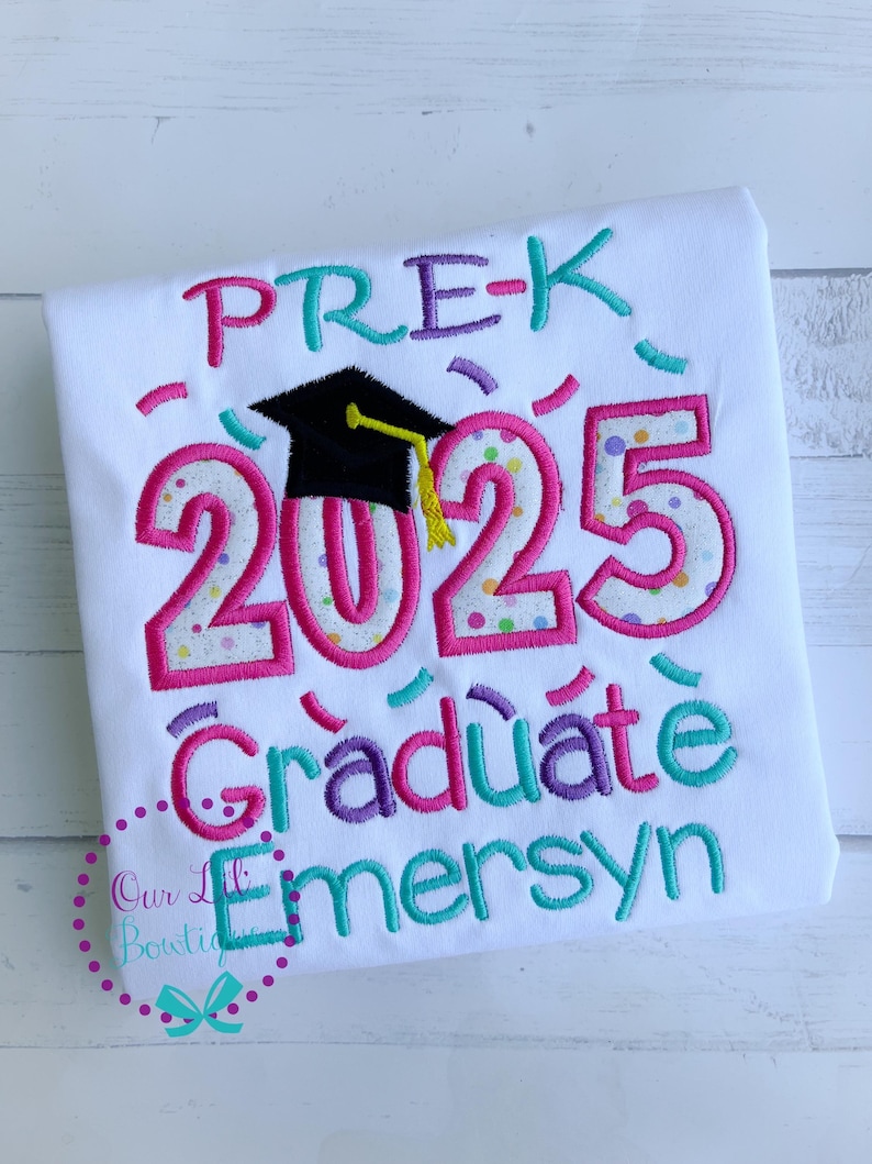 May include: White t-shirt with pink, blue, and purple embroidery that says "Pre-K 2025 Graduate" with a black graduation cap and the name "Emersyn" underneath.