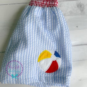 Puede incluir: Pantalones cortos de seersucker azul claro y blanco con cintura el&aacute;stica de cuadros vichy rojos y blancos. Un aplique bordado de una pelota de playa en amarillo, blanco, rojo y azul se encuentra en la parte inferior derecha. El texto Our Lil Boutique es visible.