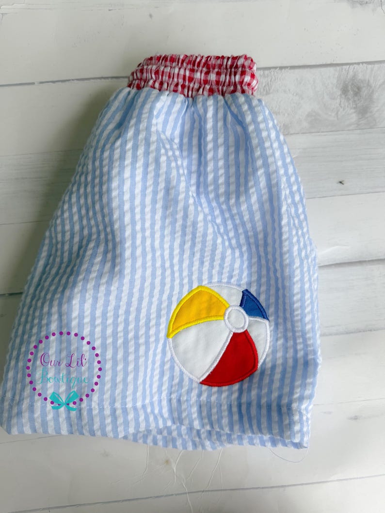 Puede incluir: Pantalones cortos de seersucker azul claro y blanco con cintura el&aacute;stica de cuadros vichy rojos y blancos. Un aplique bordado de una pelota de playa en amarillo, blanco, rojo y azul se encuentra en la parte inferior derecha. El texto Our Lil Boutique es visible.