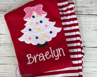 May include: A red long-sleeve shirt featuring an embroidered white Christmas tree with colorful polka dots and a pink bow on top, with the name Braelyn stitched in white below. The shirt is paired with red and white horizontally striped pants.