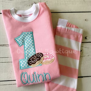May include: Pink and white striped pajamas with a pink shirt featuring a turquoise number one with white polka dots, two donuts, and the name "Quinn" embroidered in turquoise.