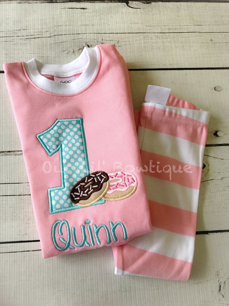 May include: Pink and white striped pajamas with a pink shirt featuring a turquoise number one with white polka dots, two donuts, and the name "Quinn" embroidered in turquoise.
