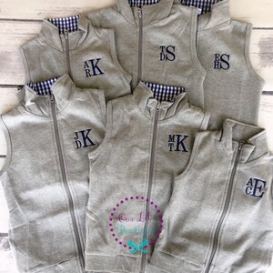May include: A collection of gray zip-up vests with navy blue monogrammed initials. The vests have a navy and white gingham lining at the collar. The vests are displayed on a light wood surface.