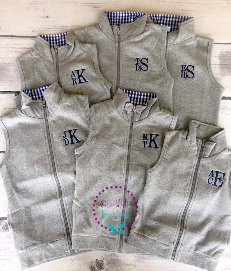 May include: A collection of gray zip-up vests with navy blue monogrammed initials. The vests have a navy and white gingham lining at the collar. The vests are displayed on a light wood surface.