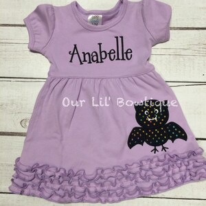 Puede incluir: Un vestido de manga corta color violeta claro con dobladillo con volantes. El vestido presenta el nombre Anabelle bordado en letra negra y una aplicaci&oacute;n de murci&eacute;lago negro con lunares de colores. Una etiqueta en el cuello indica la talla 12M.