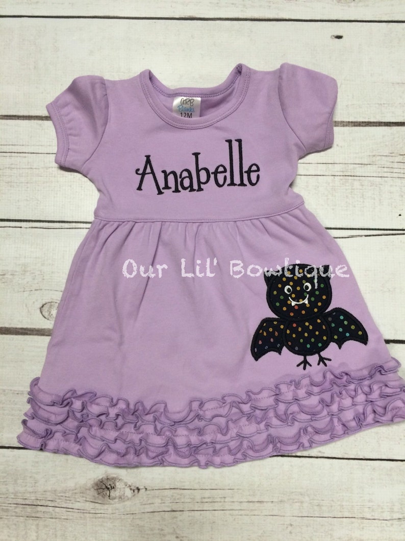 Puede incluir: Un vestido de manga corta color violeta claro con dobladillo con volantes. El vestido presenta el nombre Anabelle bordado en letra negra y una aplicaci&oacute;n de murci&eacute;lago negro con lunares de colores. Una etiqueta en el cuello indica la talla 12M.