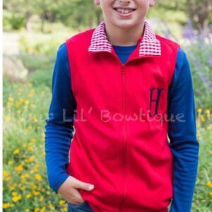 May include: A young person wearing a red zip-up vest with a blue long-sleeved shirt underneath and blue jeans. The vest has a white and red checkered collar and a navy blue initial. The text "Lil' Bowtique" is visible.