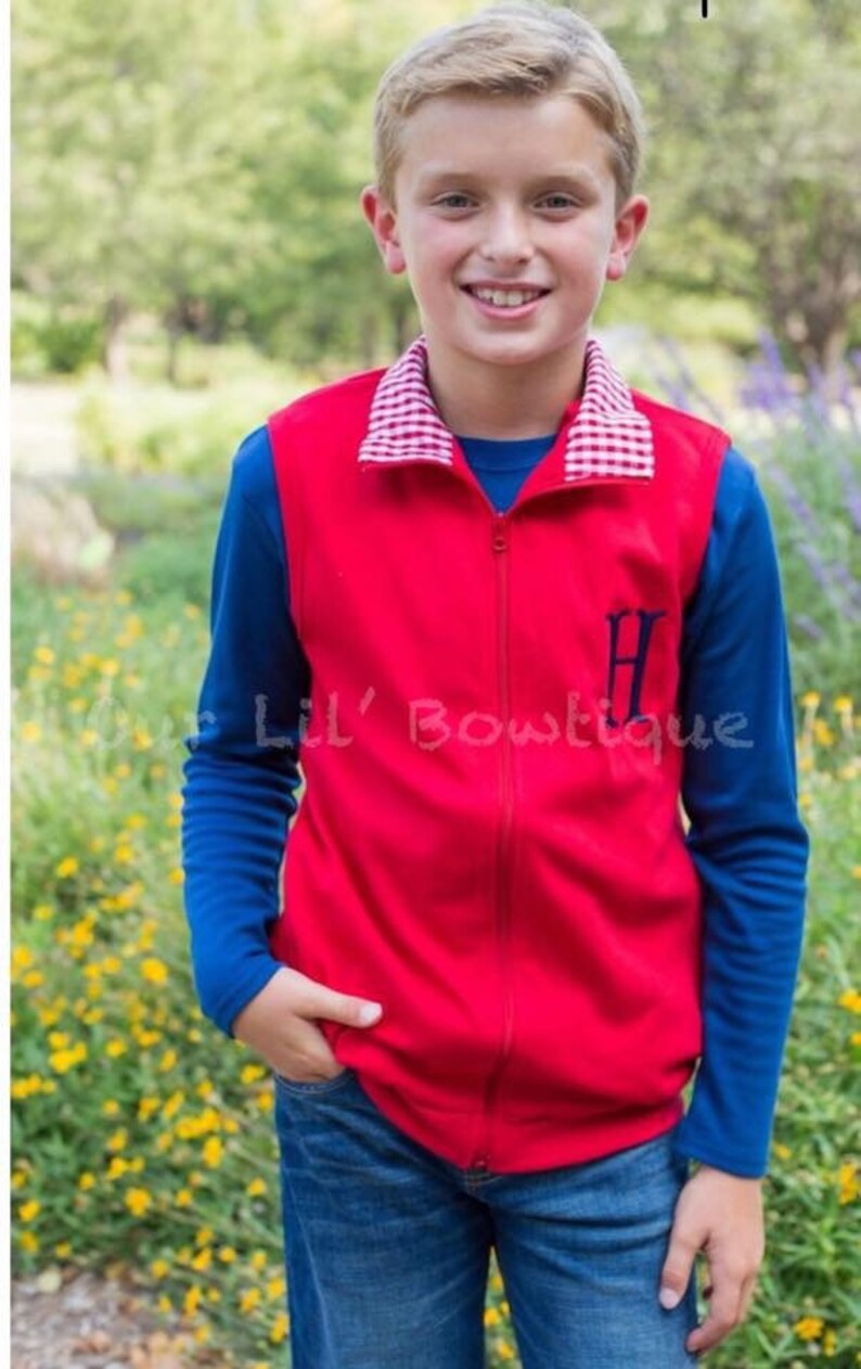May include: A young person wearing a red zip-up vest with a blue long-sleeved shirt underneath and blue jeans. The vest has a white and red checkered collar and a navy blue initial. The text "Lil' Bowtique" is visible.