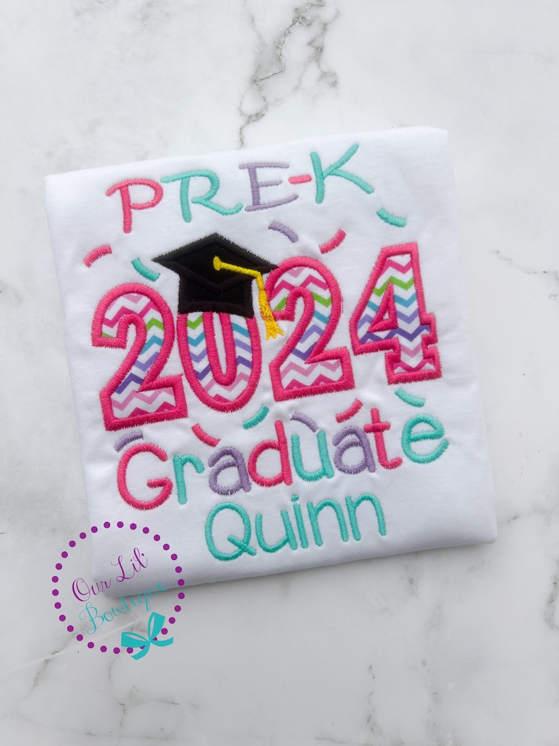 May include: White t-shirt with colorful embroidered text that says "PRE-K 2024 Graduate Quinn". The numbers are decorated with a chevron pattern and a black graduation cap with a yellow tassel is above the numbers.