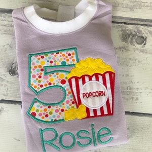 May include: Purple t-shirt with a colorful number 5 and a popcorn bucket with the word "POPCORN" on it. The name "Rosie" is embroidered below the design.