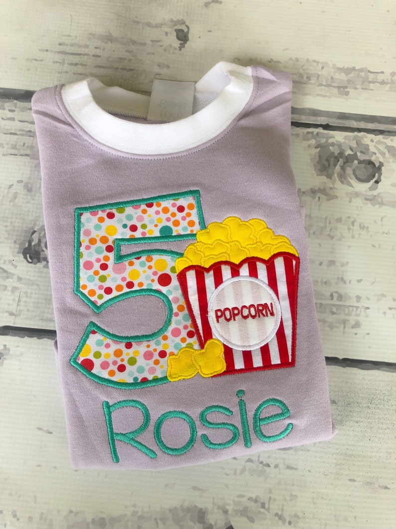 May include: Purple t-shirt with a colorful number 5 and a popcorn bucket with the word "POPCORN" on it. The name "Rosie" is embroidered below the design.