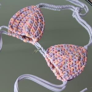 Pode incluir: Um top de biquíni em crochê com duas copas triangulares feitas de fio matizado em tons de lavanda, pêssego e rosa claro. A peça inclui laços trançados em violeta claro para o pescoço e as costas, com um pequeno pingente de cruz prateado.