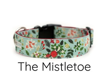 May include: A dog collar with a floral pattern in shades of blue, red, and white. The collar has a black buckle and a silver metal ring. The text "The Mistletoe" is displayed below the collar.