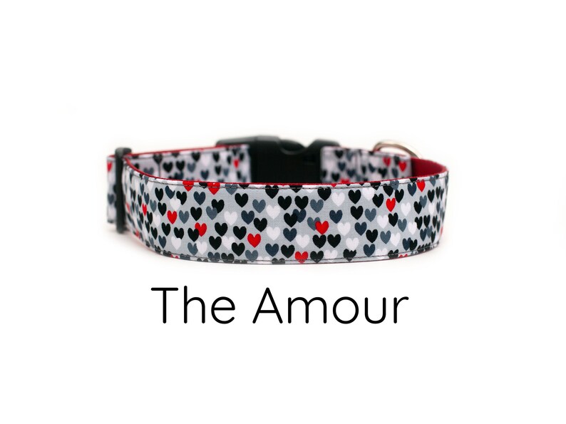 May include: A white dog collar with a red border and a pattern of small black and red hearts. The collar has a black buckle and the text "The Amour" is printed below the collar.