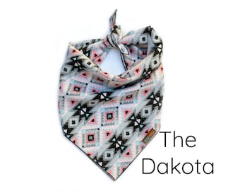 May include: A grey, black, pink and blue patterned bandana with a geometric design. The bandana is tied in a knot at the top and says "The Dakota" on the bottom right corner.