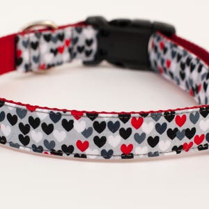 May include: A red and white dog collar with a black and gray heart pattern. The collar has a plastic buckle and a metal D-ring.