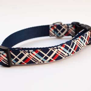 May include: A navy blue dog collar with a white, red, and blue plaid pattern. The collar has a black buckle and a silver D-ring.