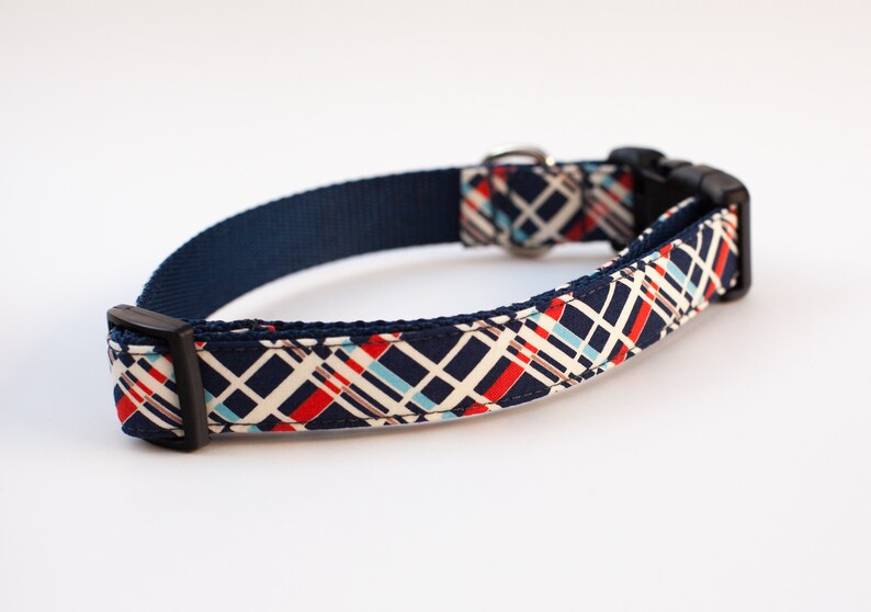 May include: A navy blue dog collar with a white, red, and blue plaid pattern. The collar has a black buckle and a silver D-ring.