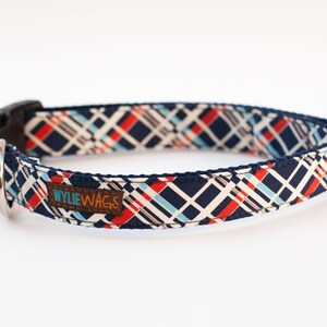 May include: A navy blue dog collar with a white, red, and blue geometric pattern. The collar has a black buckle and a silver D-ring. The collar has a brown leather tag that says "WYLIE WAGS".