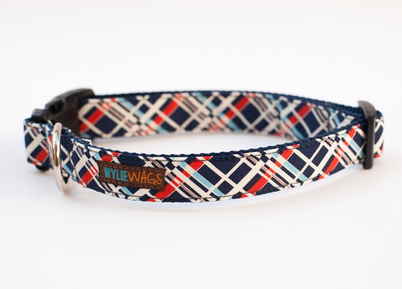 May include: A navy blue dog collar with a white, red, and blue geometric pattern. The collar has a black buckle and a silver D-ring. The collar has a brown leather tag that says "WYLIE WAGS".