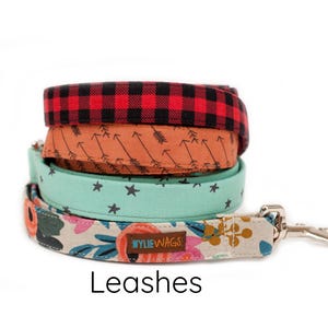 May include: A stack of four colorful dog leashes with different patterns. The leashes are made of fabric and have a metal clip. The leashes are labeled "WYLIE WAGS". Leashes.