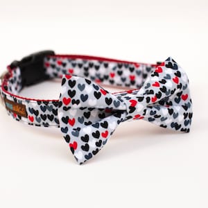 May include: A gray, white, and red dog collar with a bow tie featuring a heart pattern. The collar has the text "Pug-a-licious" on it.