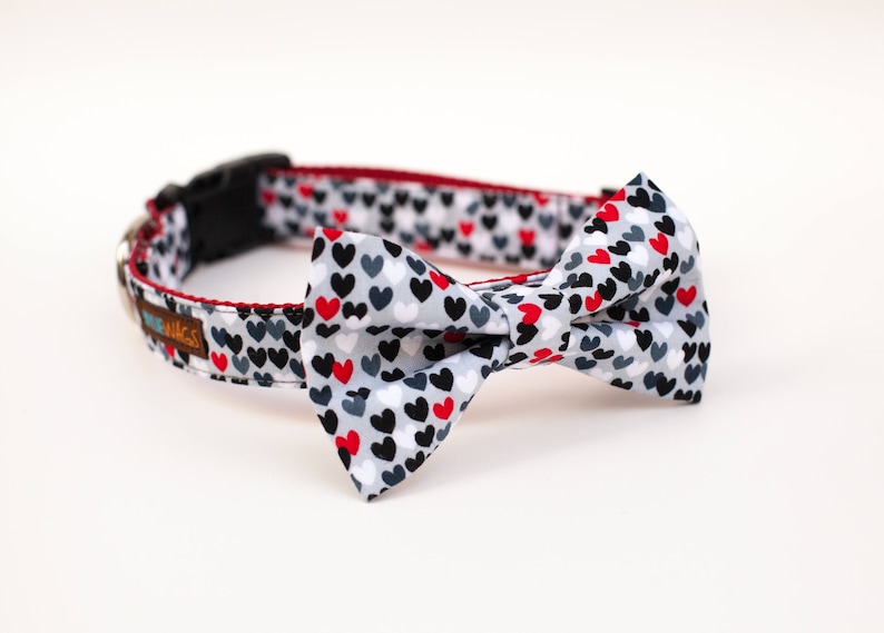 May include: A gray, white, and red dog collar with a bow tie featuring a heart pattern. The collar has the text "Pug-a-licious" on it.