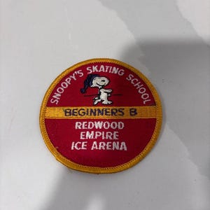 May include: A circular embroidered patch with a red background and a yellow border. It features a graphic of Snoopy wearing a blue hat, with the text SNOOPY'S SKATING SCHOOL, BEGINNERS B, and REDWOOD EMPIRE ICE ARENA embroidered in white and blue.