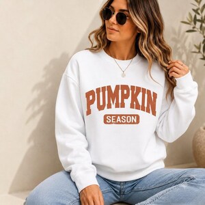 May include: A white long-sleeve crewneck sweatshirt featuring the text PUMPKIN SEASON in a distressed orange collegiate-style font. The word PUMPKIN is arched across the chest, with the word SEASON centered below it inside a rectangular box.