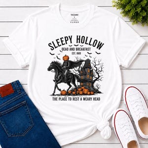 Könnte beinhalten: Ein weißes Kurzarm-T-Shirt mit einer Grafik eines kopflosen Reiters auf einem schwarzen Pferd vor einem dunklen Haus. Der Text lautet SLEEPY HOLLOW DEAD AND BREAKFAST EST. 1869 und THE PLACE TO REST A WEARY HEAD, ergänzt durch orange Kürbisse.