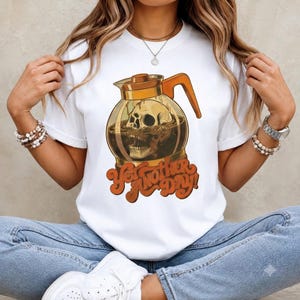 May include: A white short-sleeve t-shirt featuring a graphic of a glass coffee pot containing a human skull. Below the coffee pot, the text reads "Yet Another Day!" in a bold, orange, retro-style font. The design uses shades of orange and brown.