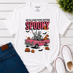 May include: A white short-sleeve t-shirt featuring a graphic of two skeletons riding in a pink convertible car. The text reads GET IN, LOSER WE'RE GETTING SPOOKY. The design includes orange pumpkins, black bats, and small star shapes surrounding the car.