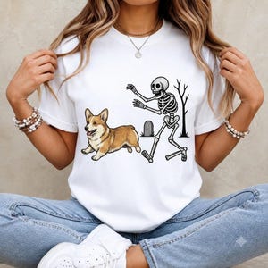 May include: A white short-sleeved t-shirt featuring a graphic print of a brown and white Corgi dog running away from a human skeleton. The skeleton is depicted with arms outstretched near a small tombstone and a bare tree silhouette.