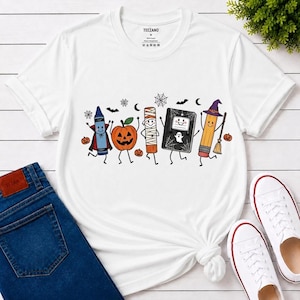 May include: A white short-sleeve t-shirt featuring a graphic print of five anthropomorphic school supplies dressed for Halloween, including a crayon vampire, a jack-o'-lantern, a mummy, a ghost-covered notebook, and a pencil with a witch hat.