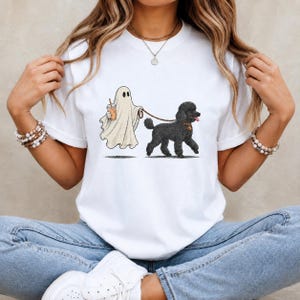 May include: A white short-sleeved t-shirt featuring a graphic print of a white ghost holding an iced coffee drink while walking a black poodle on a leash. The ghost is depicted in a classic sheet style, and the poodle has a brown collar with a tag.