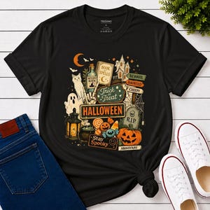 May include: A black short-sleeved t-shirt featuring a colorful Halloween-themed graphic print. The design includes a haunted house, a ghost, a cat, a pumpkin, a potion bottle, a book of spells, and signs reading Trick or Treat, Halloween, and Stay Spooky.