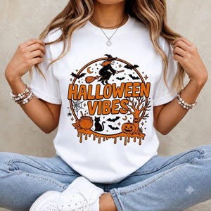 Peut inclure: Un t-shirt blanc à manches courtes avec un motif circulaire et le texte orange HALLOWEEN VIBES. Le dessin représente une sorcière sur un balai, un chat noir, un chaudron, une citrouille, un arbre dénudé, des chauves-souris et une toile.