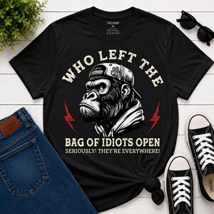 May include: A black short-sleeved t-shirt featuring a graphic of a gorilla wearing a baseball cap. The text reads: WHO LEFT THE BAG OF IDIOTS OPEN SERIOUSLY! THEY'RE EVERYWHERE! The design includes two red lightning bolt icons on the sides.