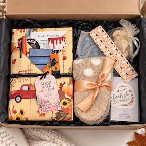 Puede incluir: Una caja de regalo con un libro envuelto en papel de temática otoñal con un camión rojo y calabazas, una etiqueta que dice Blind Date with a Book, calcetines beige suaves, un marcapáginas, una bolsa dorada pequeña y una tarjeta.