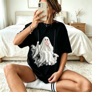Könnte beinhalten: Ein schwarzes T-Shirt mit kurzen Ärmeln und einem Grafikdruck eines weißen Geistes, der auf einer Schaukel sitzt. Der Geist trägt ein weißes Laken und bläst eine große rosa Blase mit Kaugummi. Das Motiv ist auf der Vorderseite des Shirts zentriert.