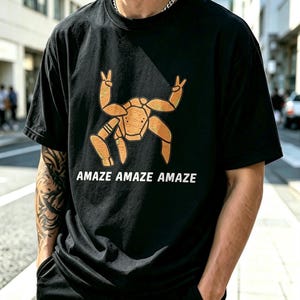 May include: A black short-sleeve t-shirt featuring a graphic of a golden-brown, multi-legged creature making peace signs with its upper limbs. Below the creature, the text "AMAZE AMAZE AMAZE" is printed in white capital letters.