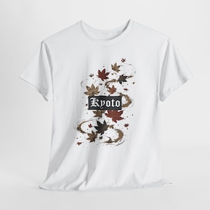 Puede incluir: Una camiseta blanca de manga corta con un gráfico central de hojas de arce dispersas en tonos rojo, marrón y negro. La palabra Kyoto está impresa en una fuente gótica blanca dentro de un recuadro rectangular negro entre las hojas.