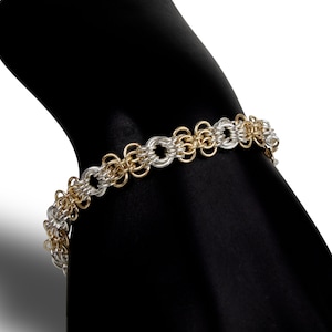 May include: A silver and gold chain bracelet with a woven design. The bracelet is on a black display stand.
