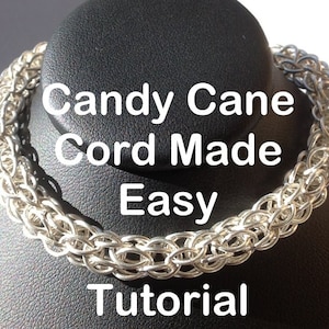 May include: A silver chain necklace made with the candy cane chainmaille pattern. The necklace is displayed on a black jewelry stand. The text "Candy Cane Cord Made Easy Tutorial" is superimposed on the image.