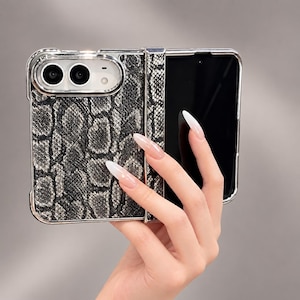 Peut inclure: Une coque de smartphone pliable avec un motif texturé en peau de serpent gris et noir. La coque présente un cadre en argent métallique et un module photo proéminent avec deux objectifs circulaires et un flash. Inclut un cache-charnière vertical.